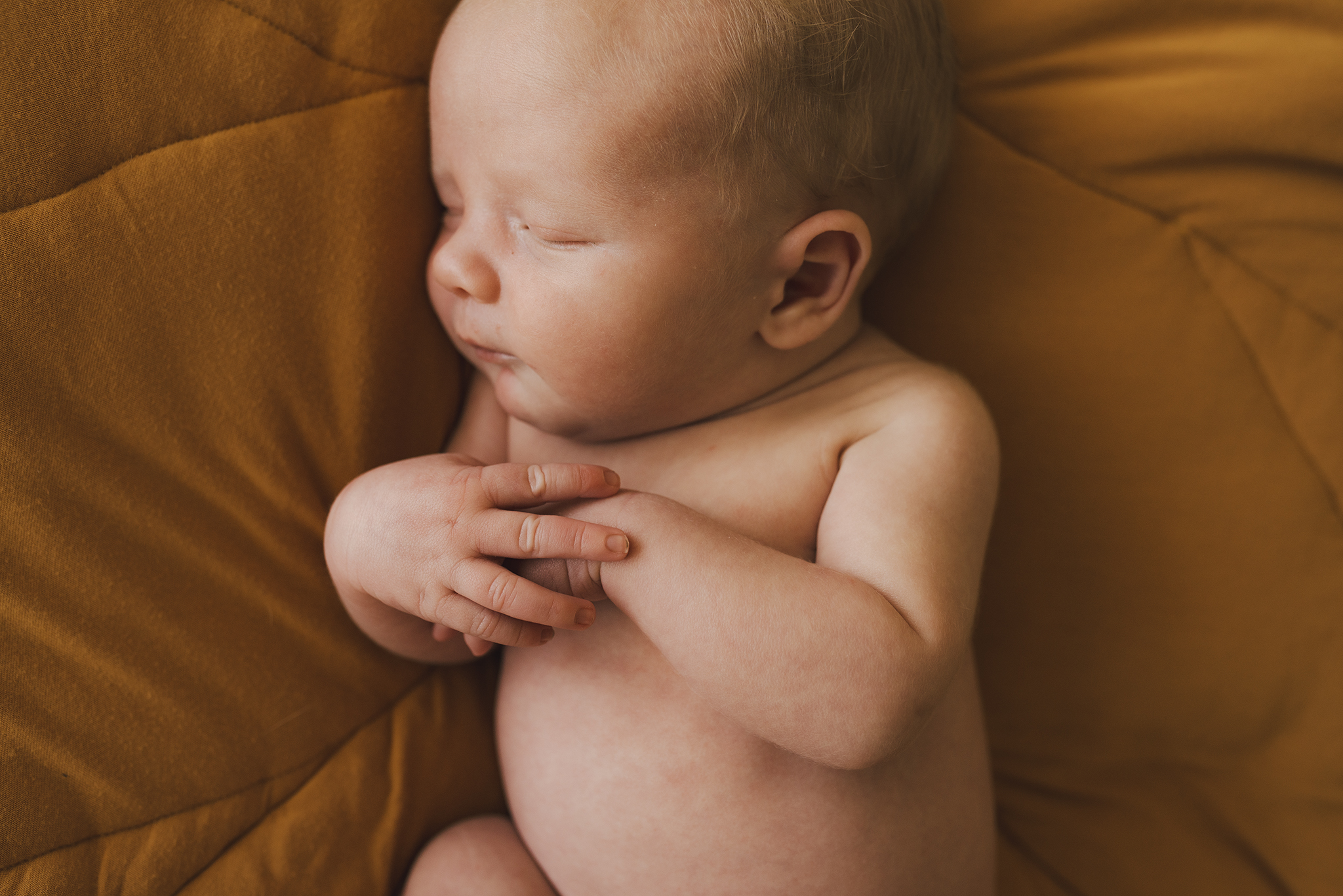 A close up of a newborn baby, laying asleep on a yellow blanket with his hands clasped together on his chest.