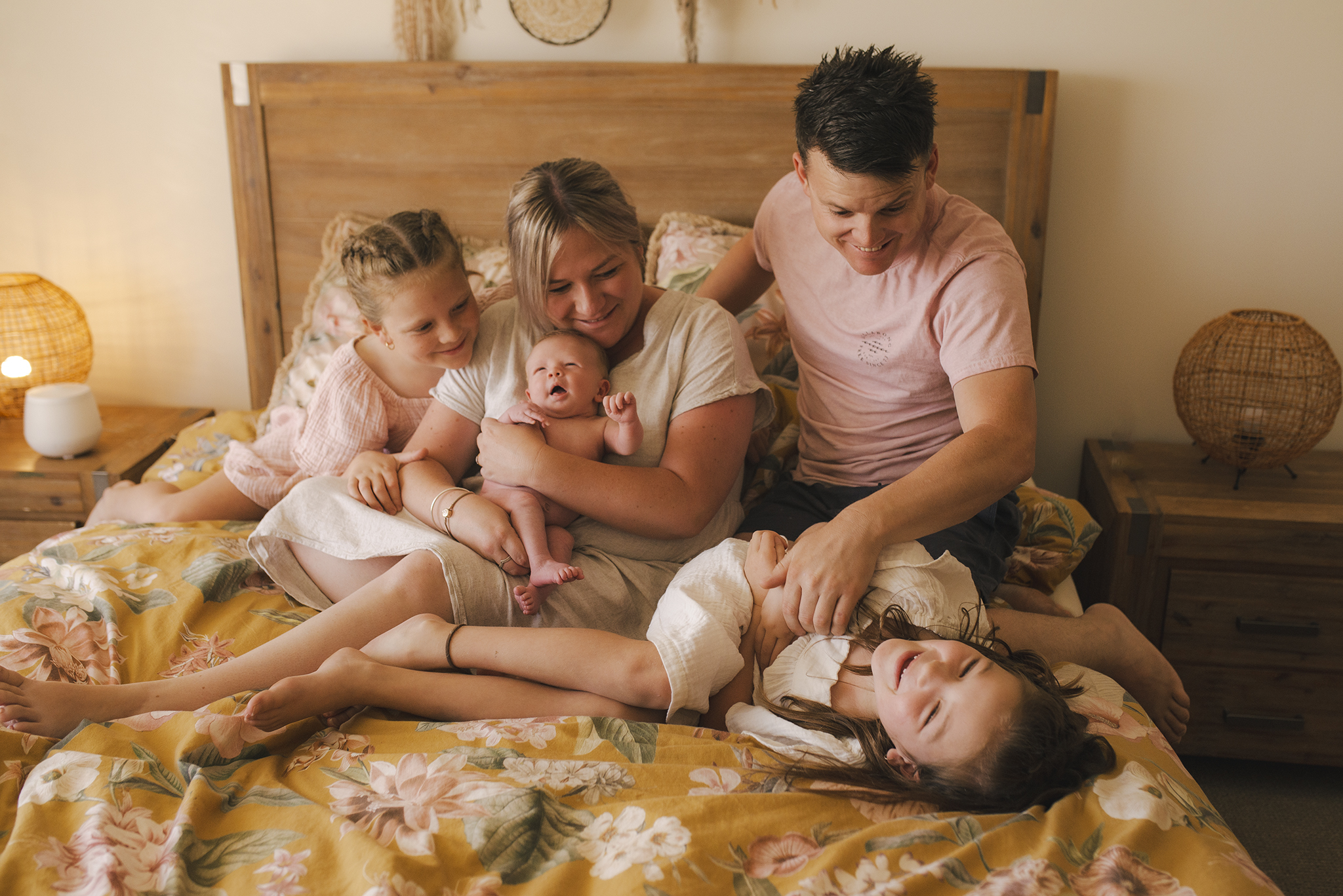 A family newborn photo. The family are sitting on a large oak timber bed with mustard & soft pink floral linen. They all sit cuddled together Mums arms around a newborn baby in the centre. One sister lays laughing at the front while dad tickles her. They are all dressed in light linens an smiling joyfully.