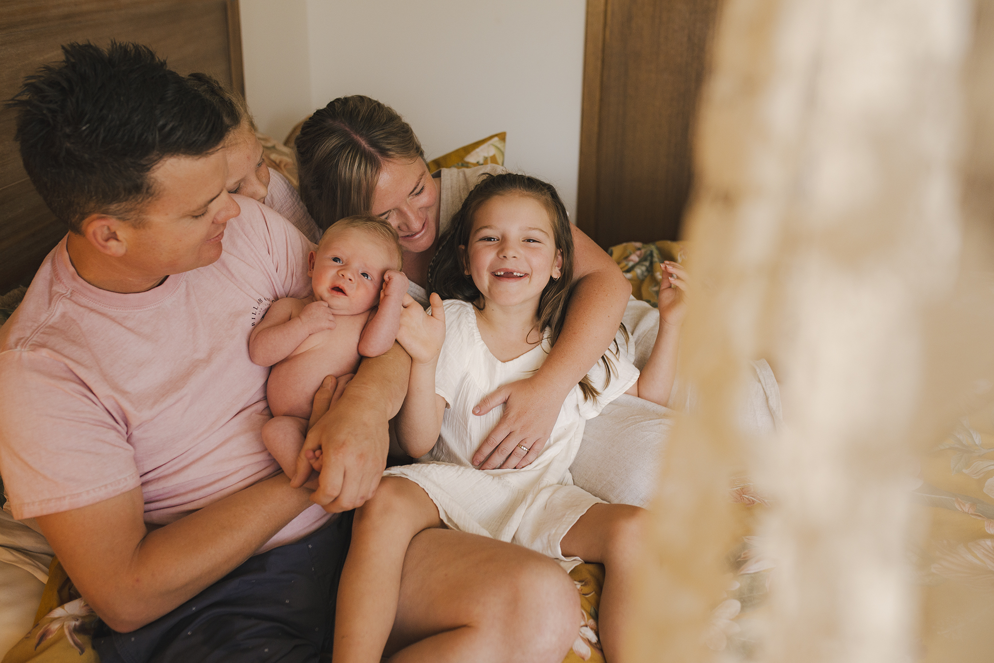 A newborn family photo. The family are snuggled up together on a large oak bed to the left of the frame with the newborn in dads arms. Everyone is smiling and interacting with each other. To the right of the frame a panel of lace curtain billows across the image giving the impression the photographer is looking into the room from outside.