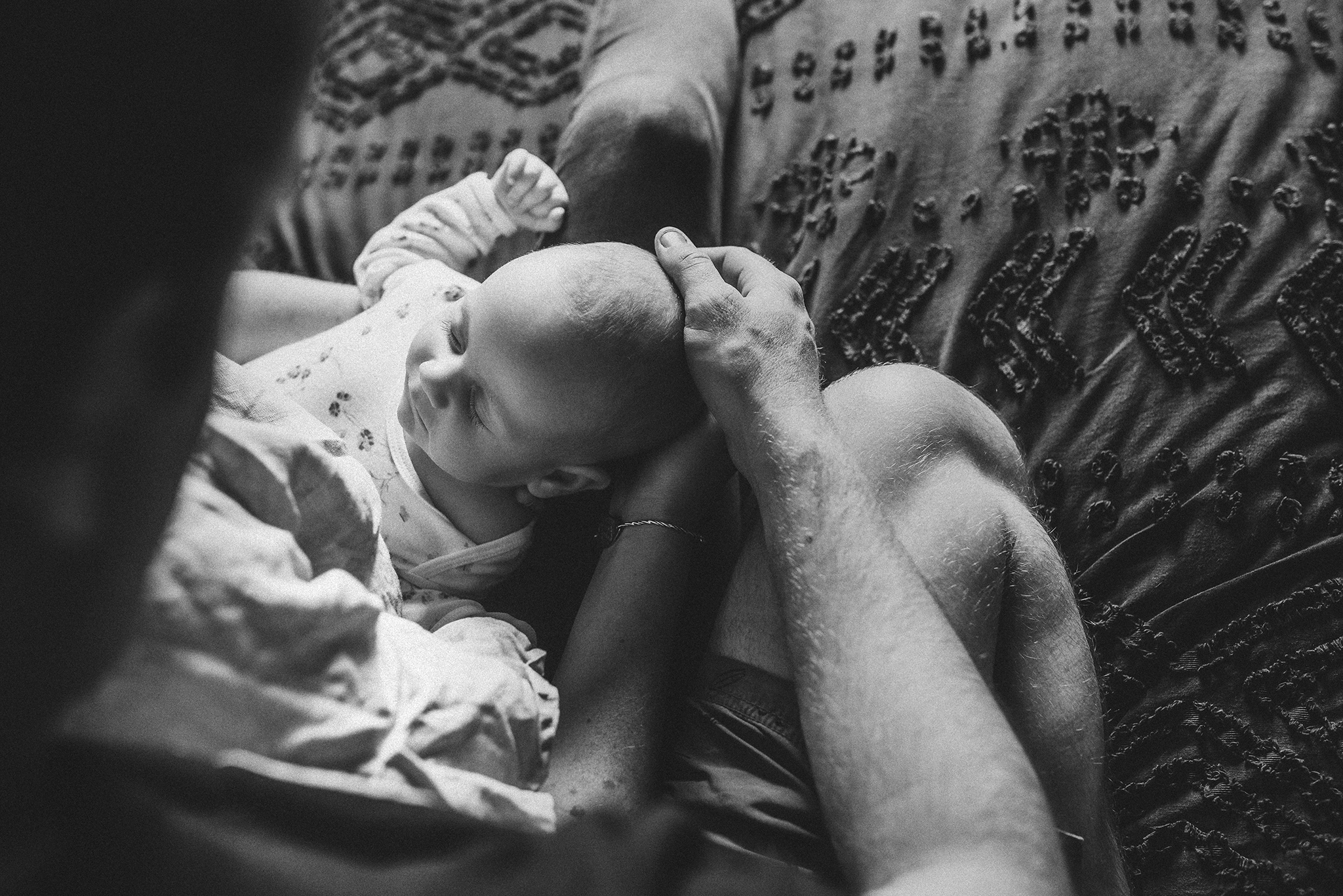 A black and white newborn image, looking over the shoulder of a mum and dad, down towards the sleeping baby cradled in their arms.