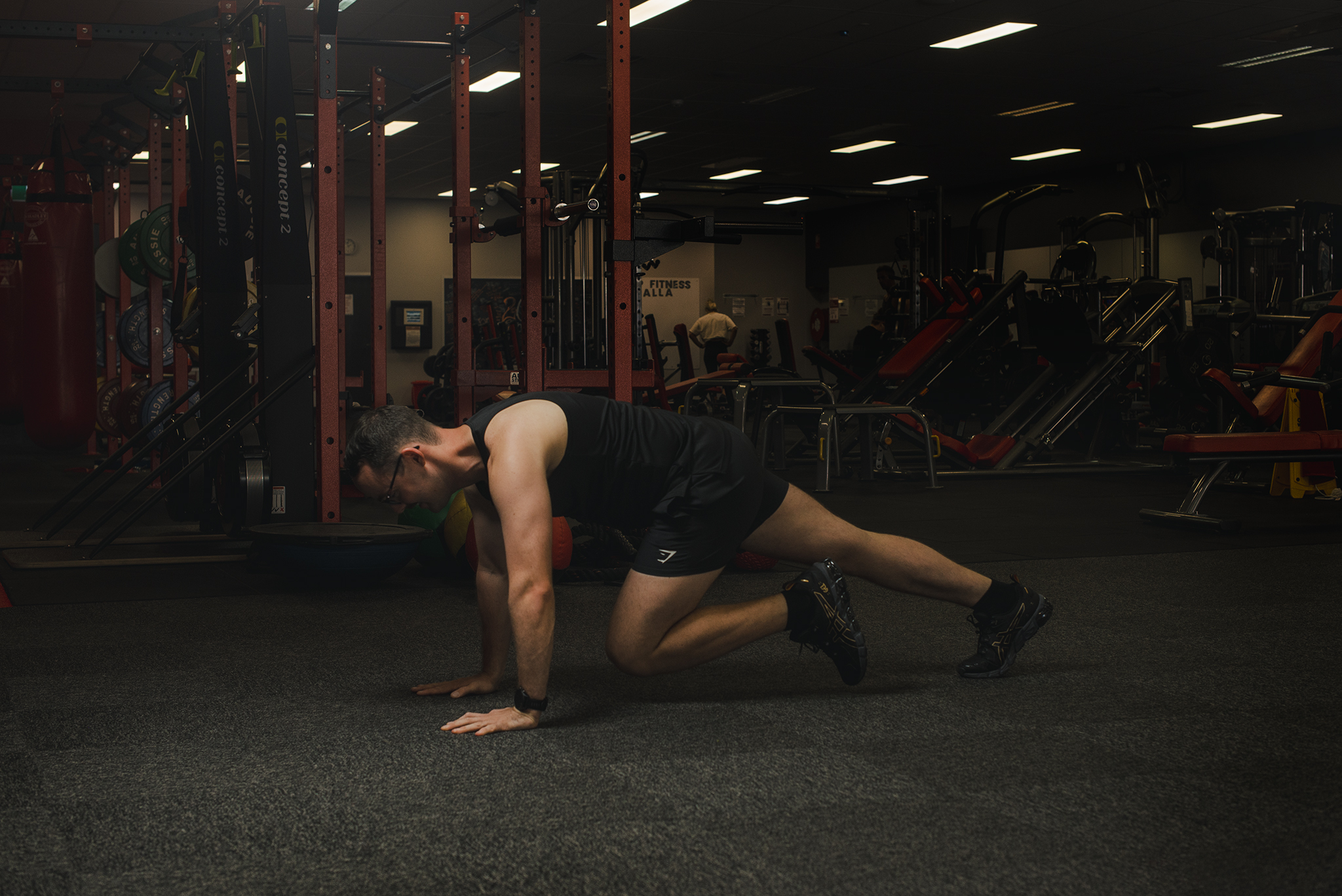 Image shows a side view of a personal trainer on all fours, in a bear crawl position with a dark and moody lit gym in the background.