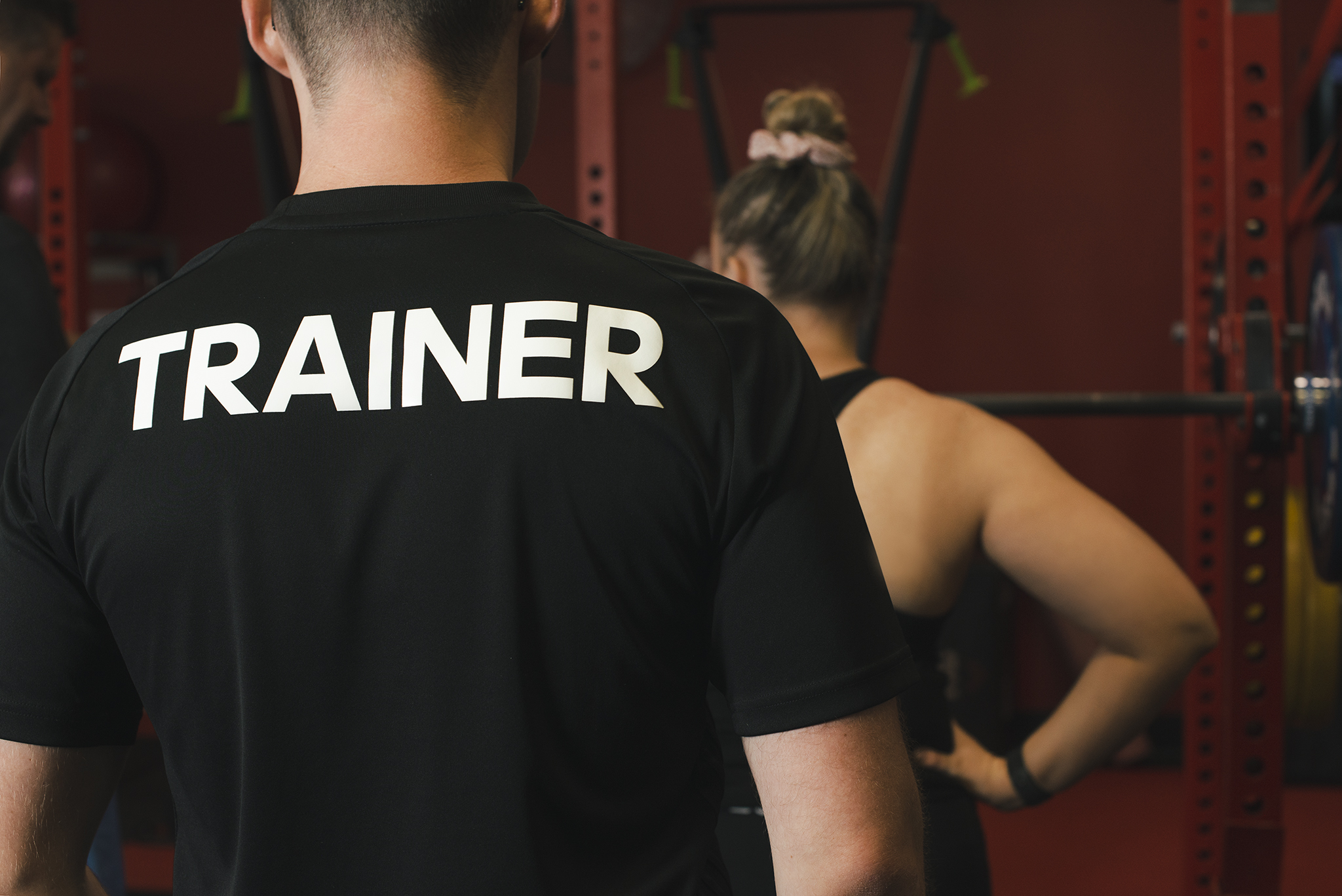 The camera looks over the shoulder of a personal trainer on the left and focuses on the word TRAINER, which is written in white on the back of a black shirt. In the background you can see a fitness client standing at a weight bar in a red gym room. The image gives the impression that the trainer is watching the client in their workout.