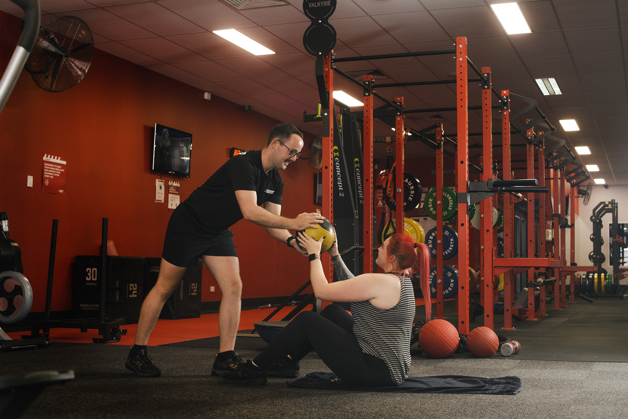 A personal trainer passes a weighted ball from a standing position down to a seated fitness client. They are in a red gym.