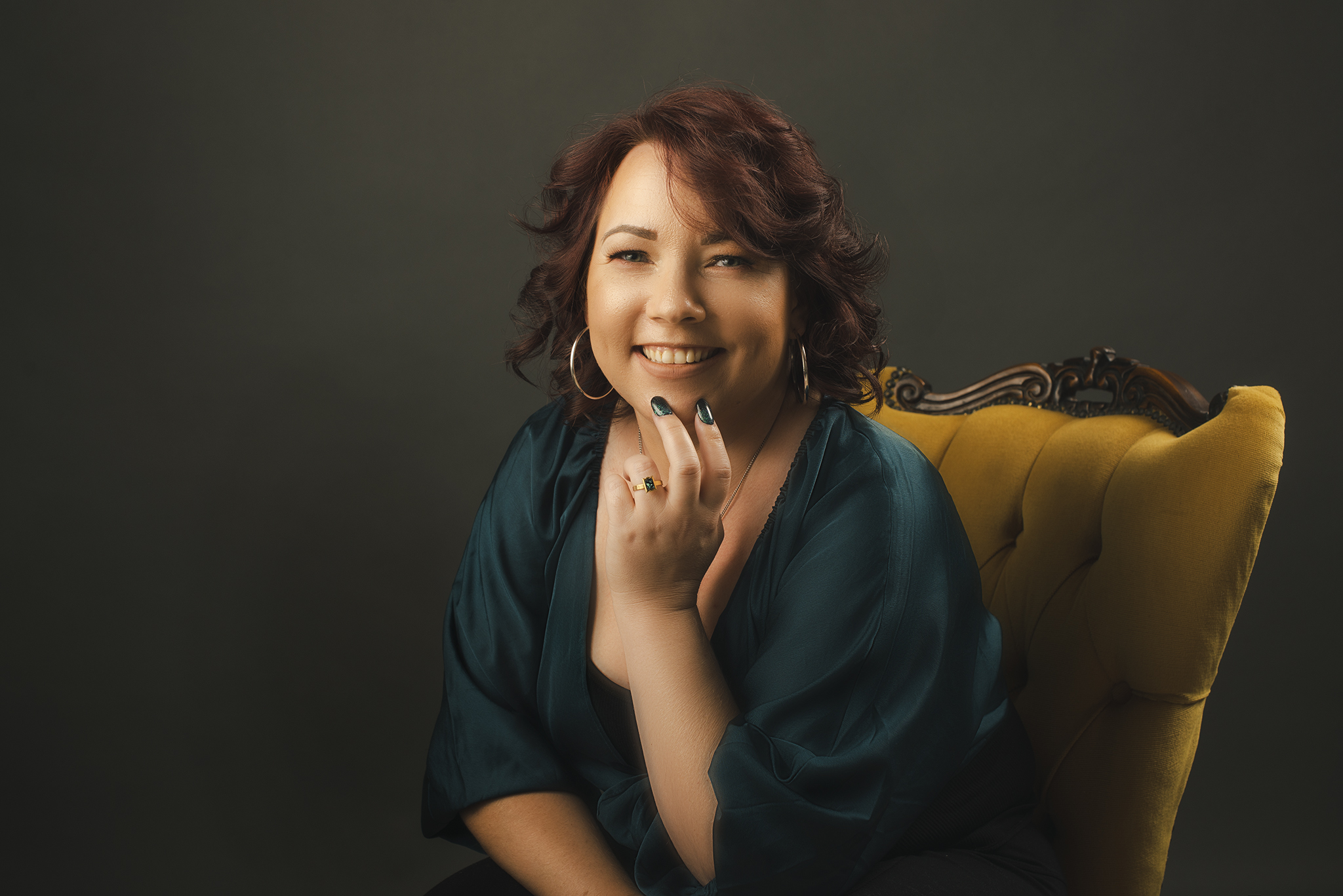 A creative headshot of a lady sitting in a dark room on a mustard, vintage, upholstered & wood carved chair. She has fiery red curls, wears large hoop earrings, an emerald satin shirt & leans towards the camera with an elegant smile. Her hand is placed up by her chin showing her emerald nails & a vintage style emerald ring on her finger.